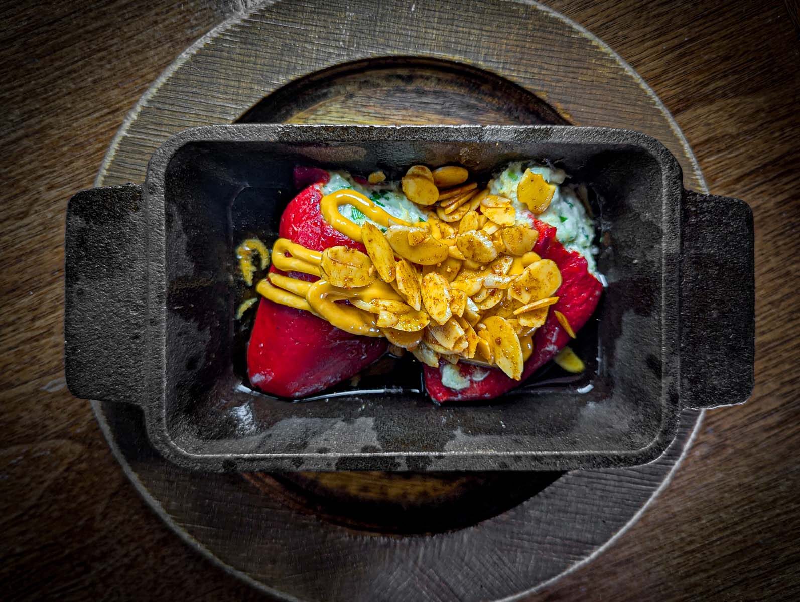 A rustic black cast iron dish holds two roasted red piquillo peppers generously stuffed with creamy crab filling. The peppers are topped with a glossy layer of toasted almond slivers and a drizzle of golden aioli. The dish sits on a round wooden charger, highlighting its artisanal presentation.
