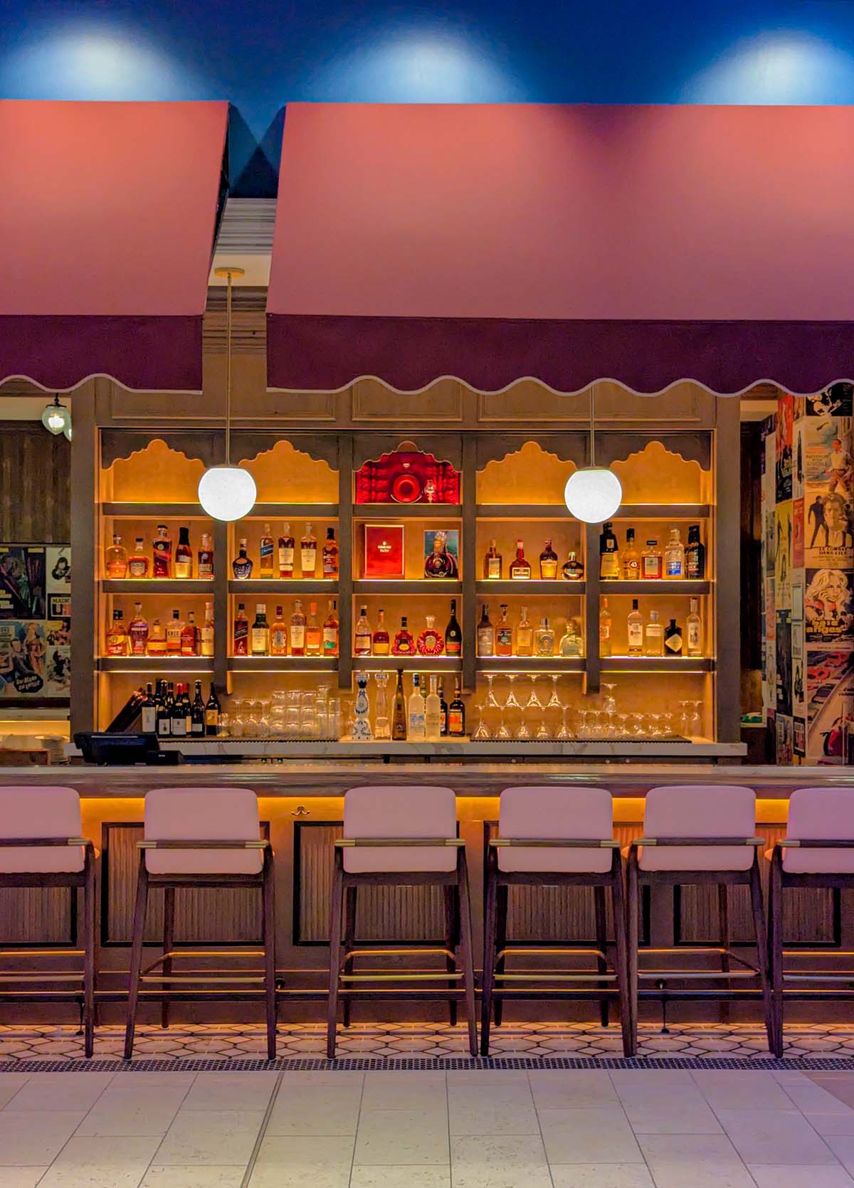 Restaurant bar with pink walls, vintage movie posters, warm lighting, and rows of liquor bottles on illuminated shelves