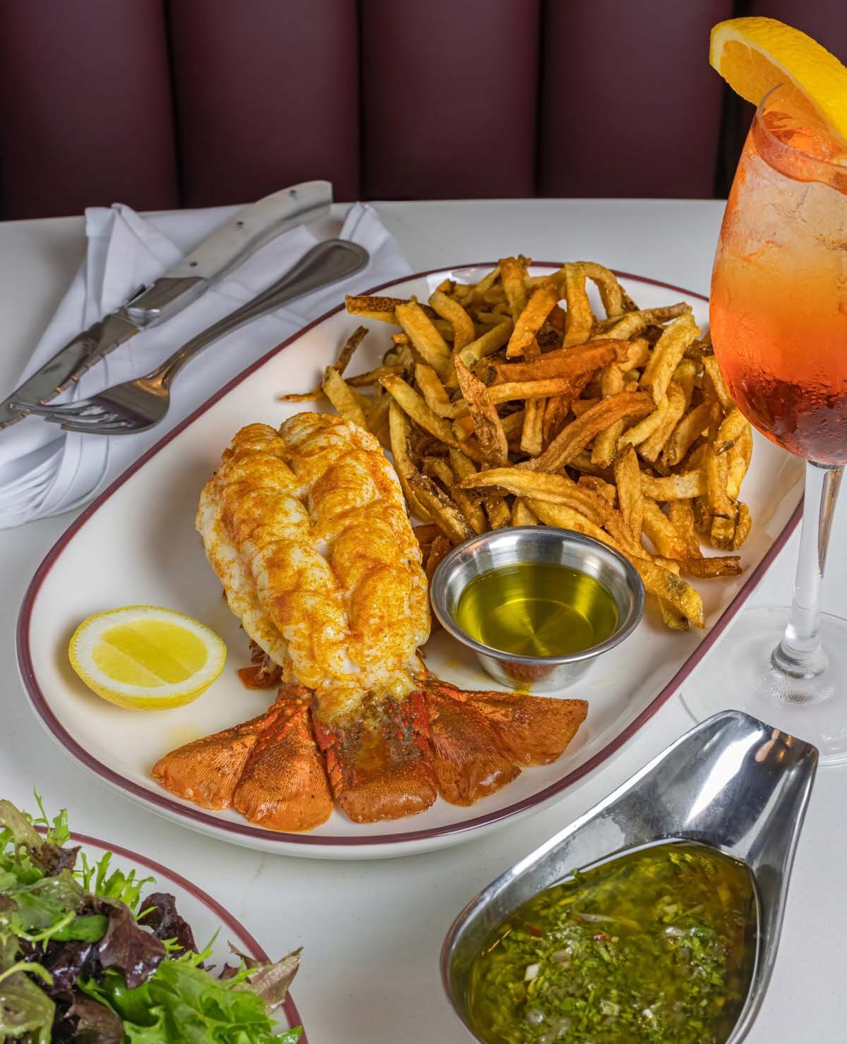 A plate with a large lobster tail split open next to golden french fries, with a small dish of melted butter and a boat of green chimichurri sauce, served on a white plate with burgundy trim