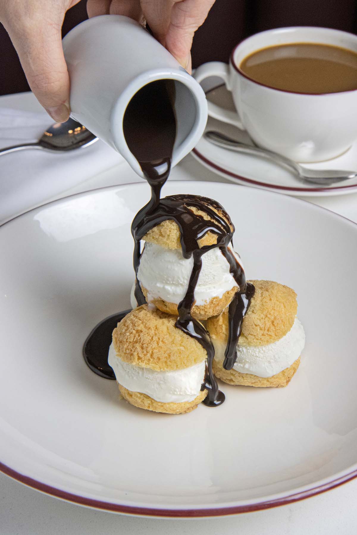 Three cream puffs filled with vanilla ice cream being drizzled with dark chocolate sauce from a white pitcher