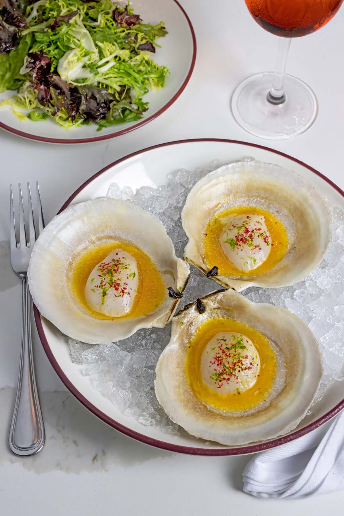 Three large scallops served in shells on ice with green herbs and red pepper garnish