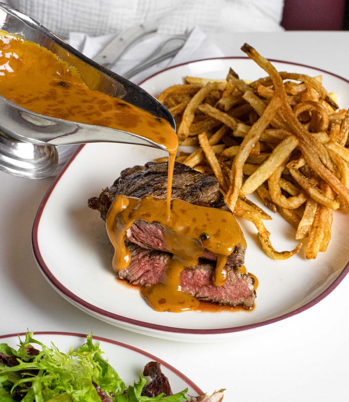 Sliced steak cooked medium-rare being topped with béarnaise sauce from a silver sauce boat, served alongside golden hand-cut french fries on a white plate with burgundy trim, with a mixed green salad visible in the foreground