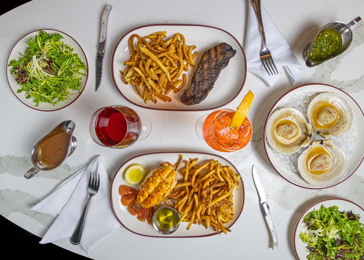 Overhead view of a table with steak frites, lobster frites, mixed green salad, cocktails, and scallops in shells
