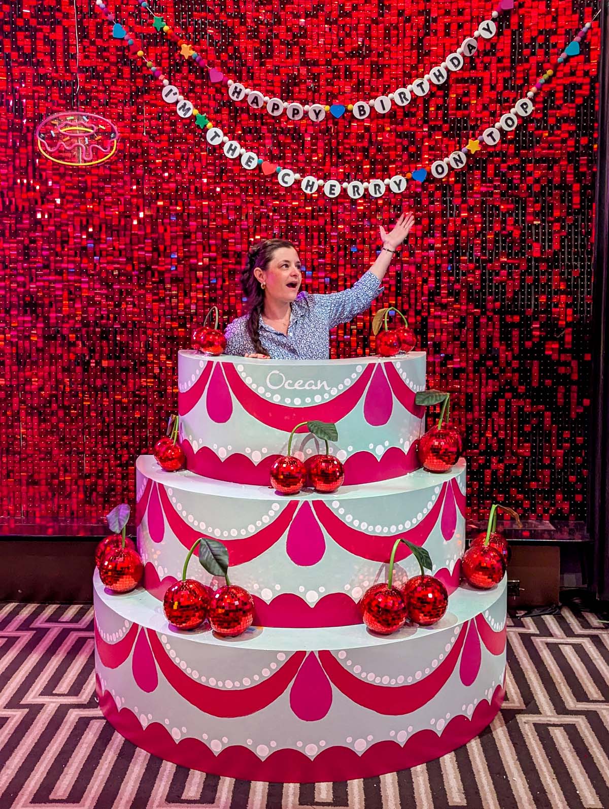 Woman popping out of giant three-tiered fake birthday cake prop with red sequined wall backdrop and cherry decorations
