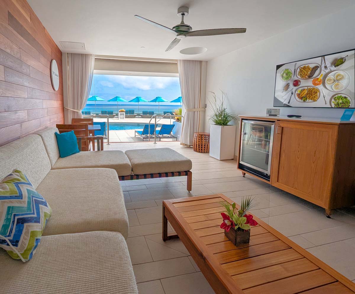 View from inside a pool cabana showing comfortable seating, mini fridge, TV, and open view to pool deck with ocean beyond