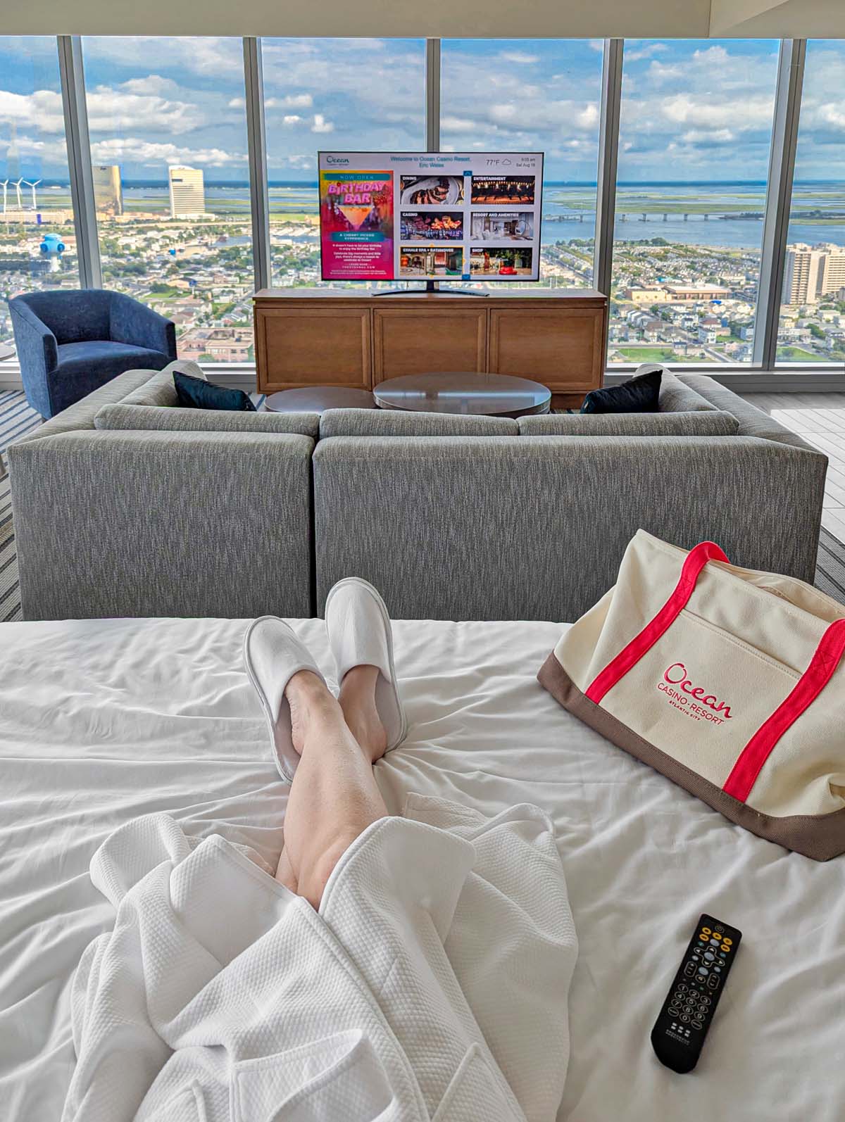 Hotel suite living room with gray sectional sofa, Ocean tote bag on white bed, floor-to-ceiling windows showing Atlantic City views