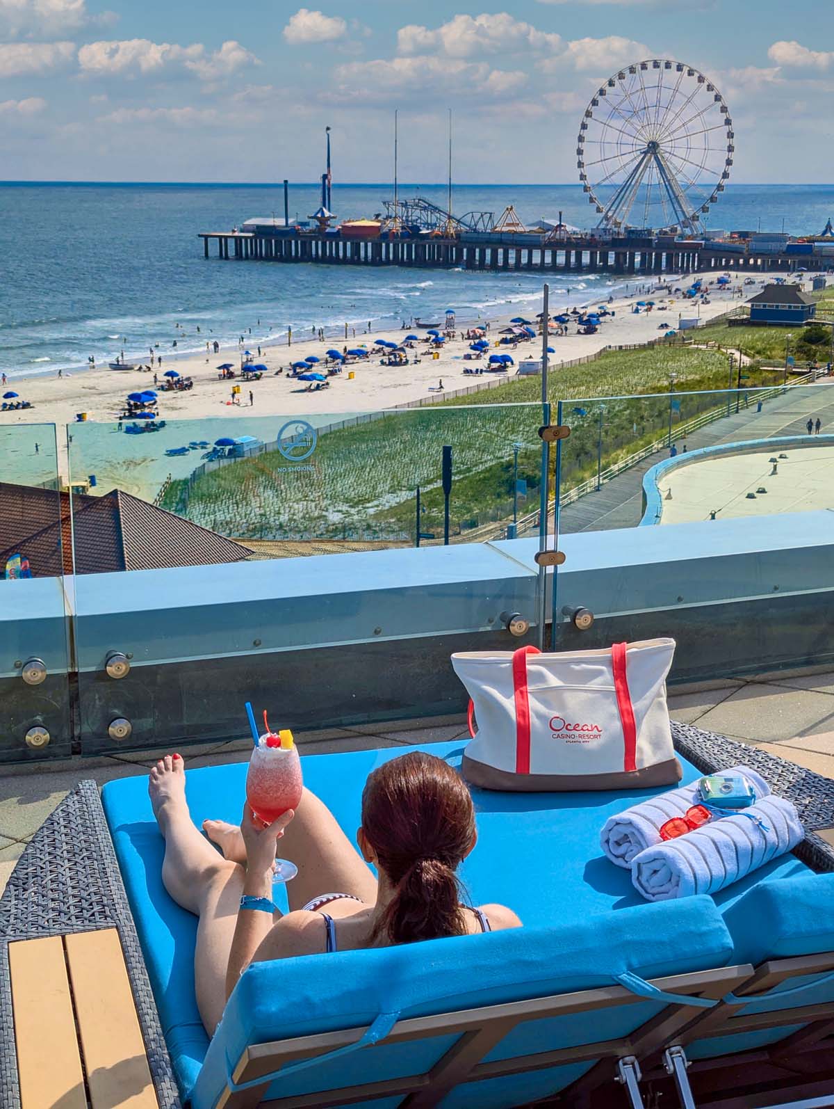 View from blue pool daybed showing Steel Pier with Ferris wheel, beach umbrellas, Ocean tote bag, and frozen cocktail