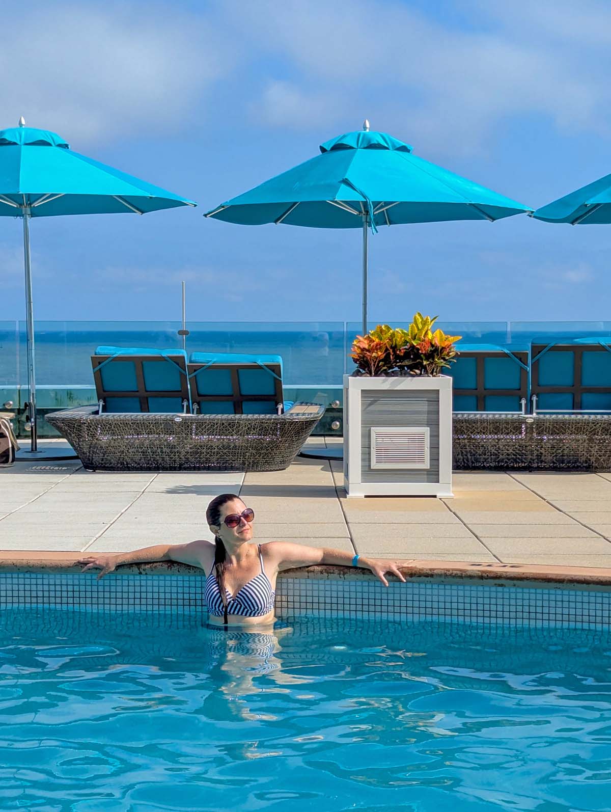 Woman in striped swimsuit relaxing in pool with turquoise umbrellas and ocean visible in background