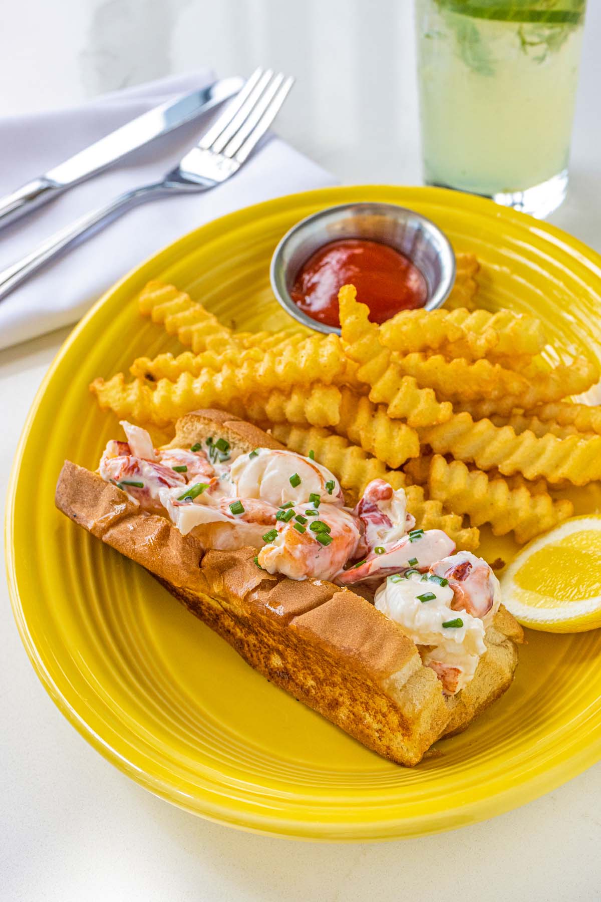 Lobster roll overflowing with chunks of lobster meat on toasted bun, served on bright yellow plate with crinkle-cut fries
