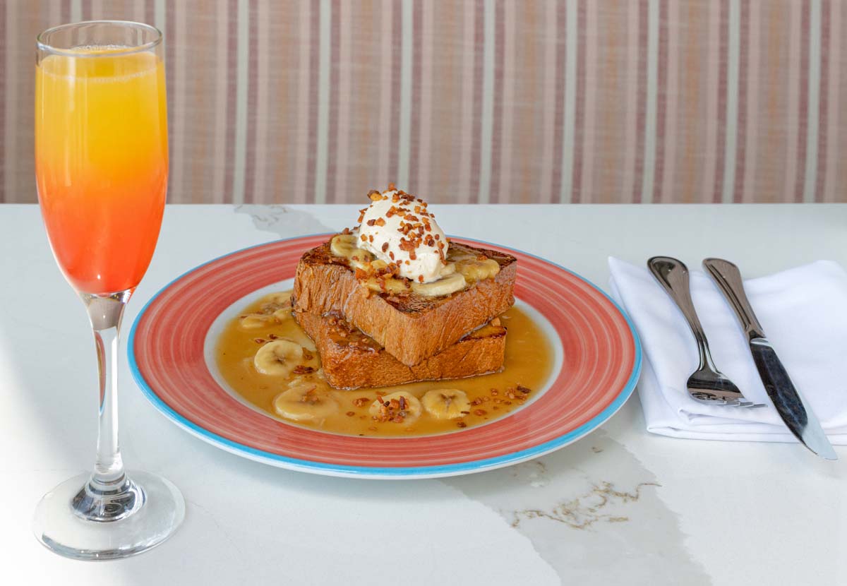 Double stack French toast topped with sliced bananas, whipped cream, and caramel sauce, with orange mimosa