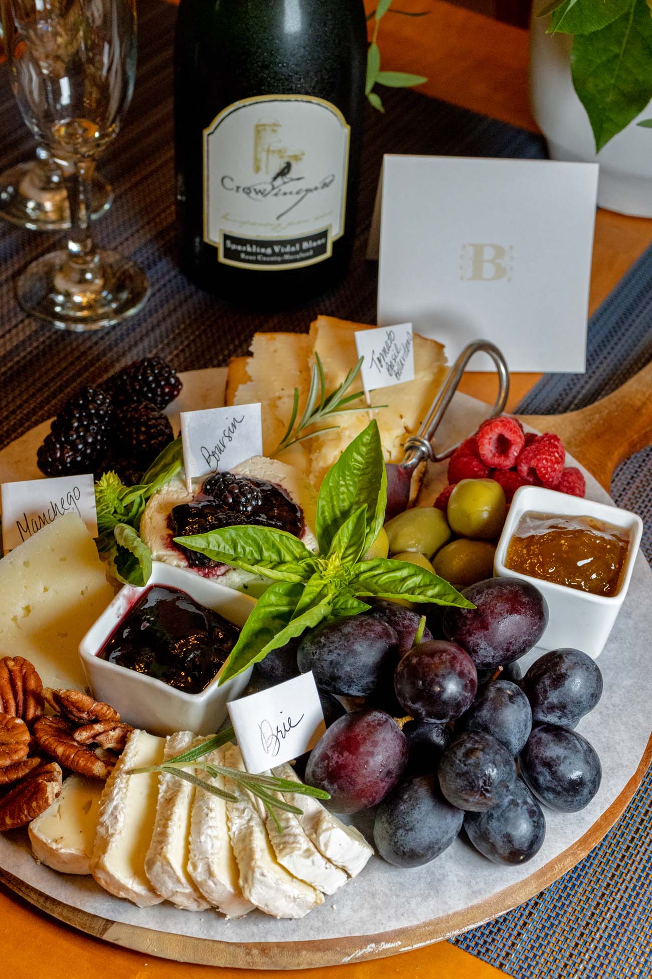 An artisan cheese and charcuterie board with Crow Sparkling Vidal Blanc wine, featuring labeled cheeses, fresh grapes, berries, and spreads arranged on a wooden platter as Brampton 1860's welcome amenity.