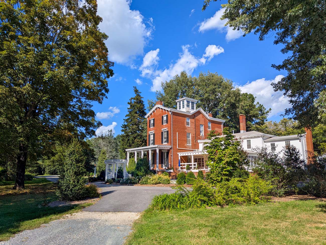 Brampton 1860's Manor House viewed from the gravel driveway approach, showing the full three-story brick facade with white trim and the property's pastoral setting.