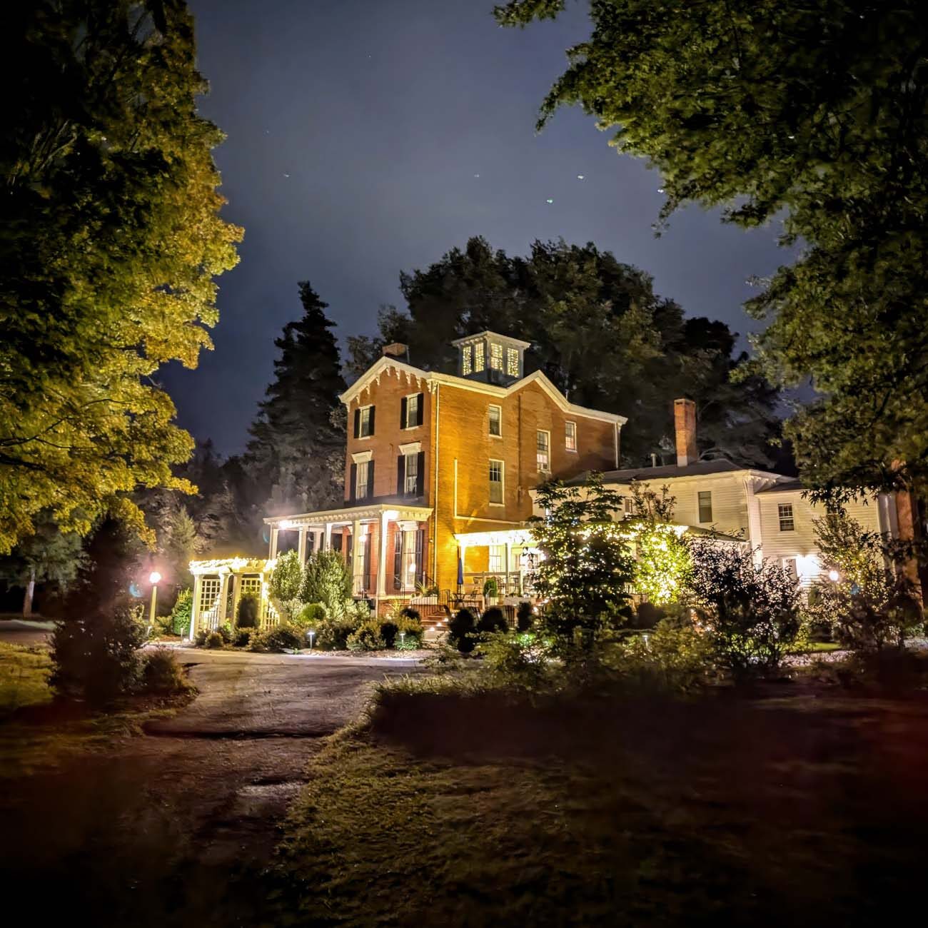 Brampton 1860's Manor House illuminated at twilight with warm golden lighting highlighting the brick facade and wraparound porch, framed by mature trees under a starlit sky.
