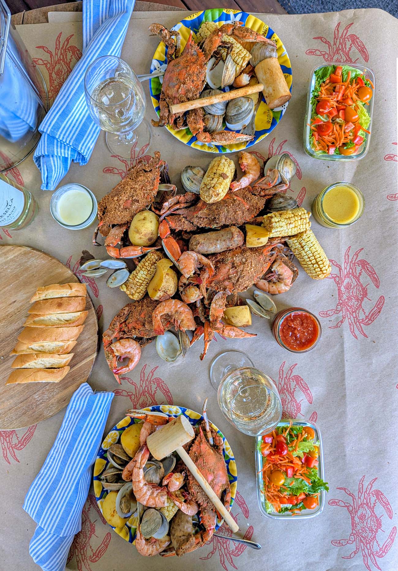 Brampton's Signature Steampot spread featuring local crabs, shrimp, clams, corn, potatoes, and kielbasa on brown paper with small salads, bread, and wine glasses.