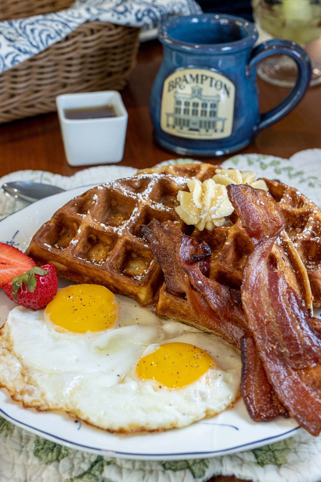 A hearty breakfast plate featuring golden waffles with butter, crispy bacon, and two sunny-side-up eggs alongside a blue ceramic Brampton 1860 commemorative mug.