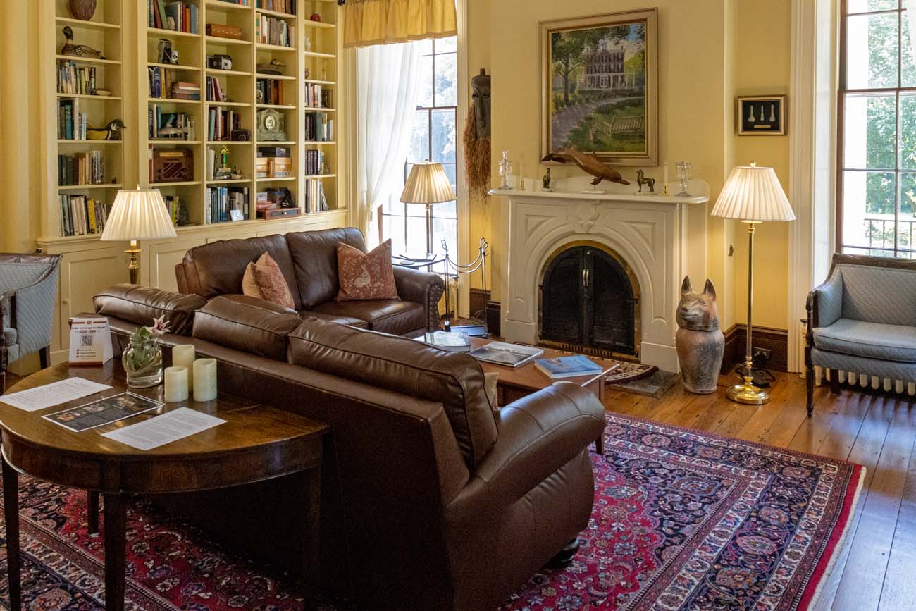 Elegant library and living room at Brampton 1860 historic inn featuring floor-to-ceiling built-in bookshelves filled with books, a large brown leather sofa with decorative pillows, traditional Persian rugs in burgundy and blue patterns, polished hardwood floors, a white marble fireplace with decorative mantel, brass table lamps with pleated shades, a round wooden coffee table with scattered magazines, tall windows with natural light, yellow walls, and a decorative ceramic cat sculpture beside the fireplace.