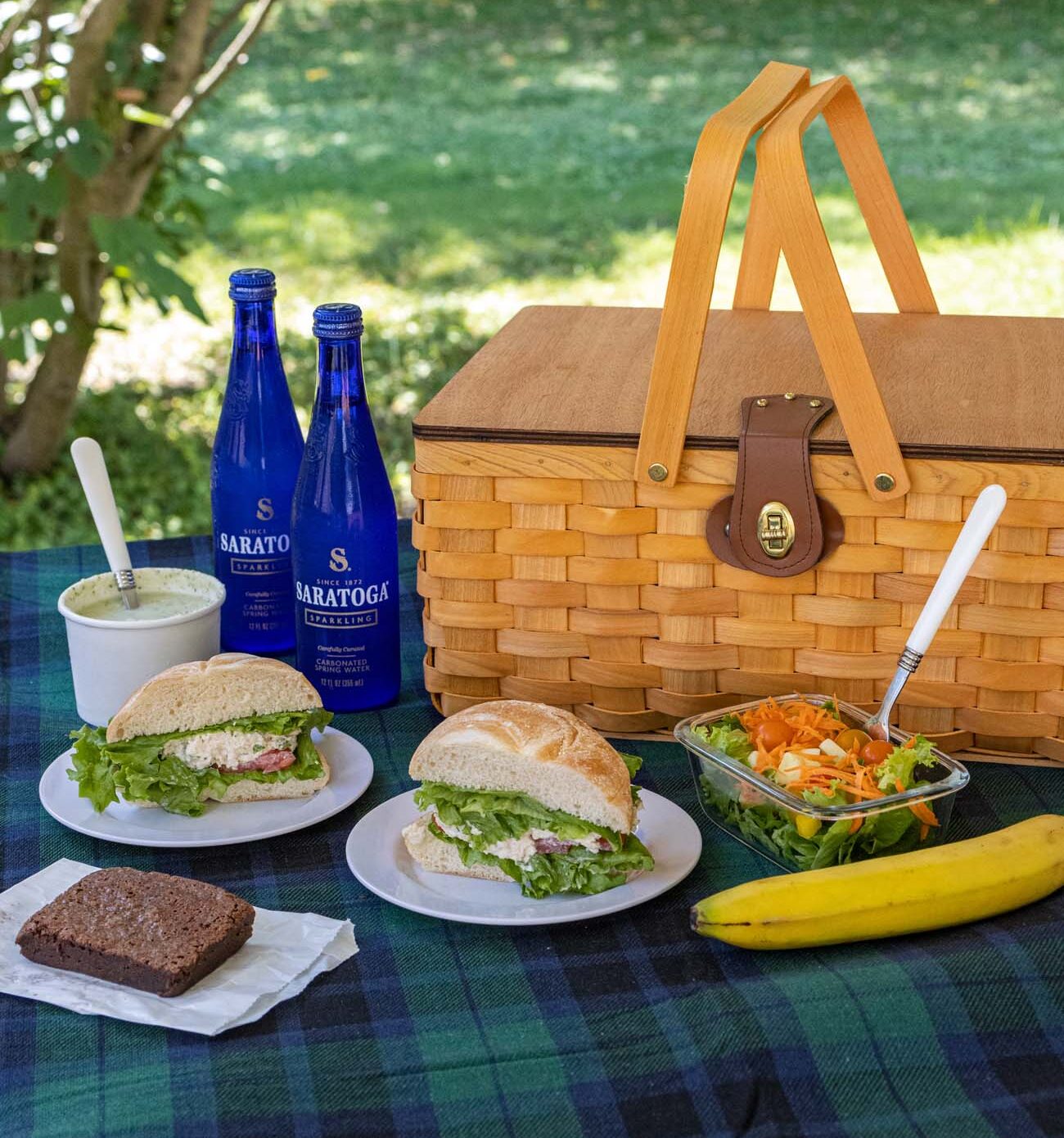 A gourmet picnic basket with Carole's chicken salad sandwiches, fresh salads, brownies, fruit, and Saratoga sparkling water arranged on a checkered blanket under trees.