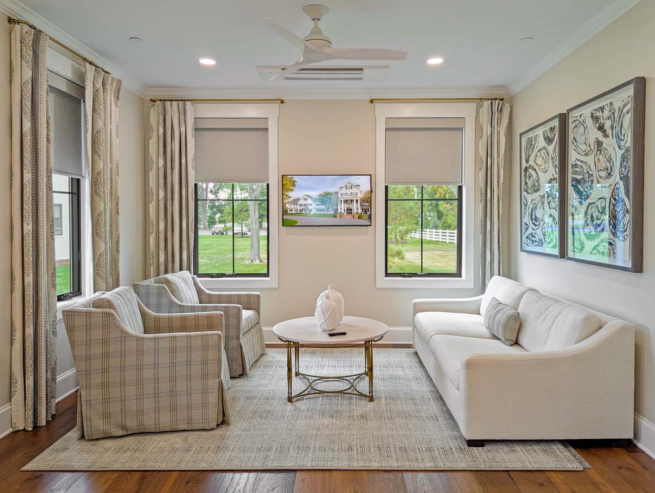 Bright, elegantly furnished cottage living room featuring cream-colored sofa and plaid upholstered armchairs arranged around a round glass coffee table. Large windows with neutral Roman shades frame views of green lawn and water beyond. The space includes warm wood floors, neutral area rug, and coastal-themed artwork on pale gray walls.