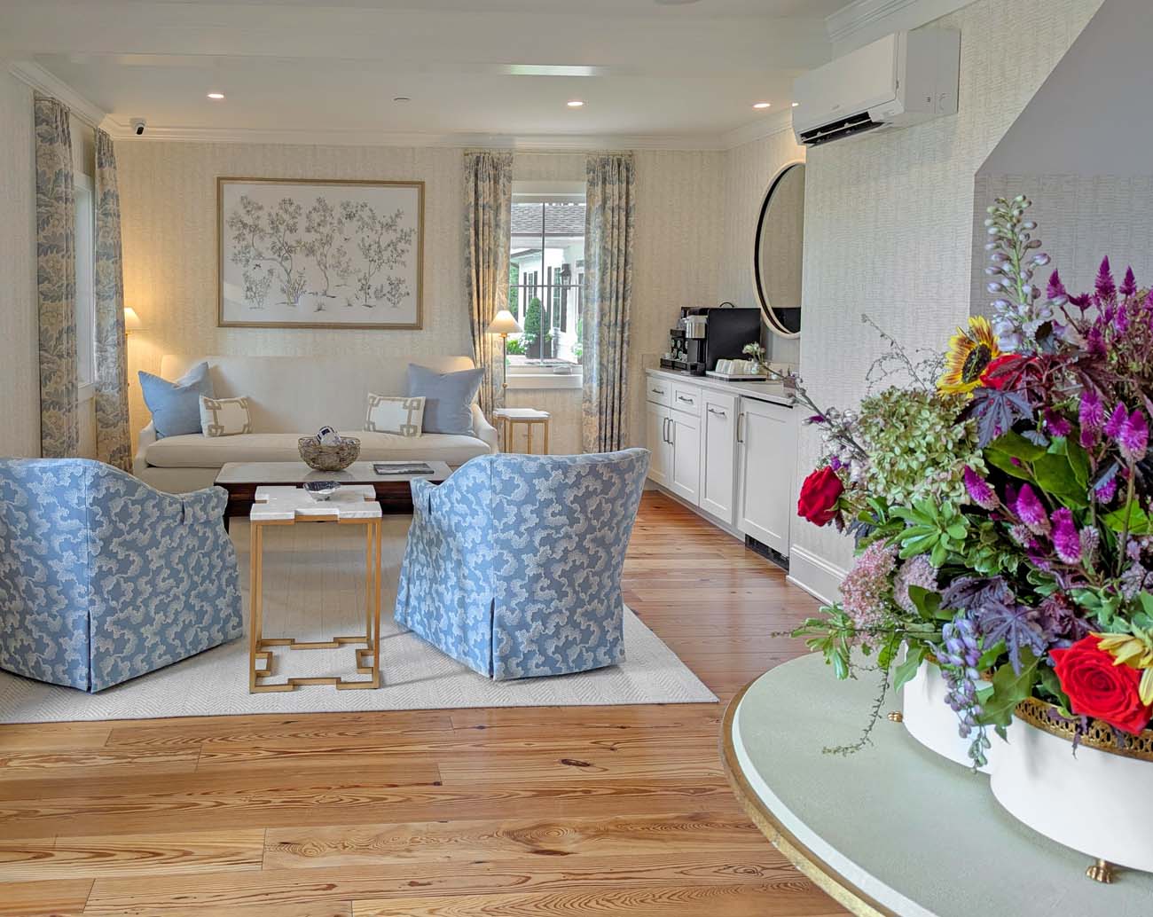 Bright, elegantly furnished lobby featuring cream-colored sofa and plaid upholstered armchairs arranged around a round glass coffee table. Large windows with neutral Roman shades frame views of green lawn and water beyond. The space includes warm wood floors, neutral area rug, and coastal-themed artwork on pale gray walls.