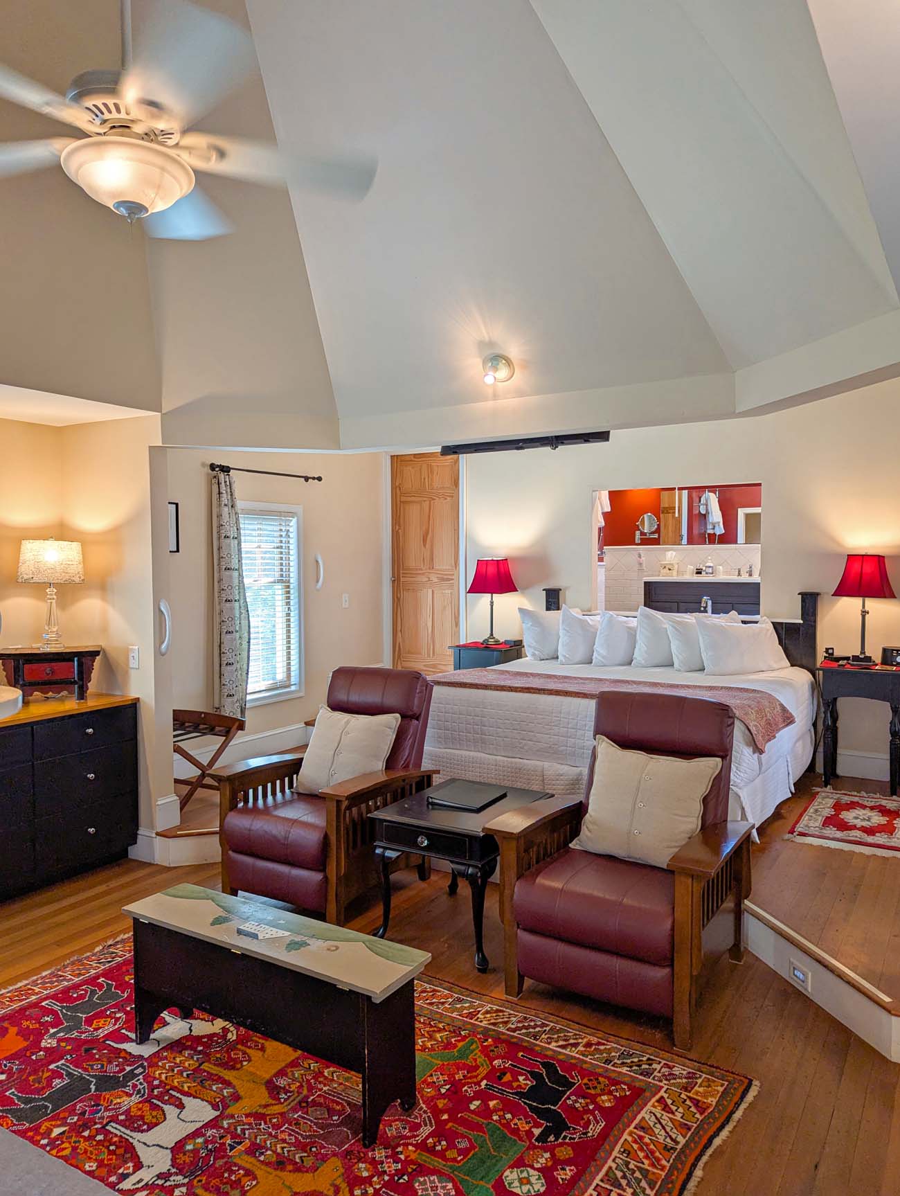 Bright, airy cottage bedroom with vaulted ceiling, king bed with white linens and burgundy leather bench at foot, two burgundy leather recliners, sliding barn door to bathroom, red accent lamps, colorful area rug, hardwood floors