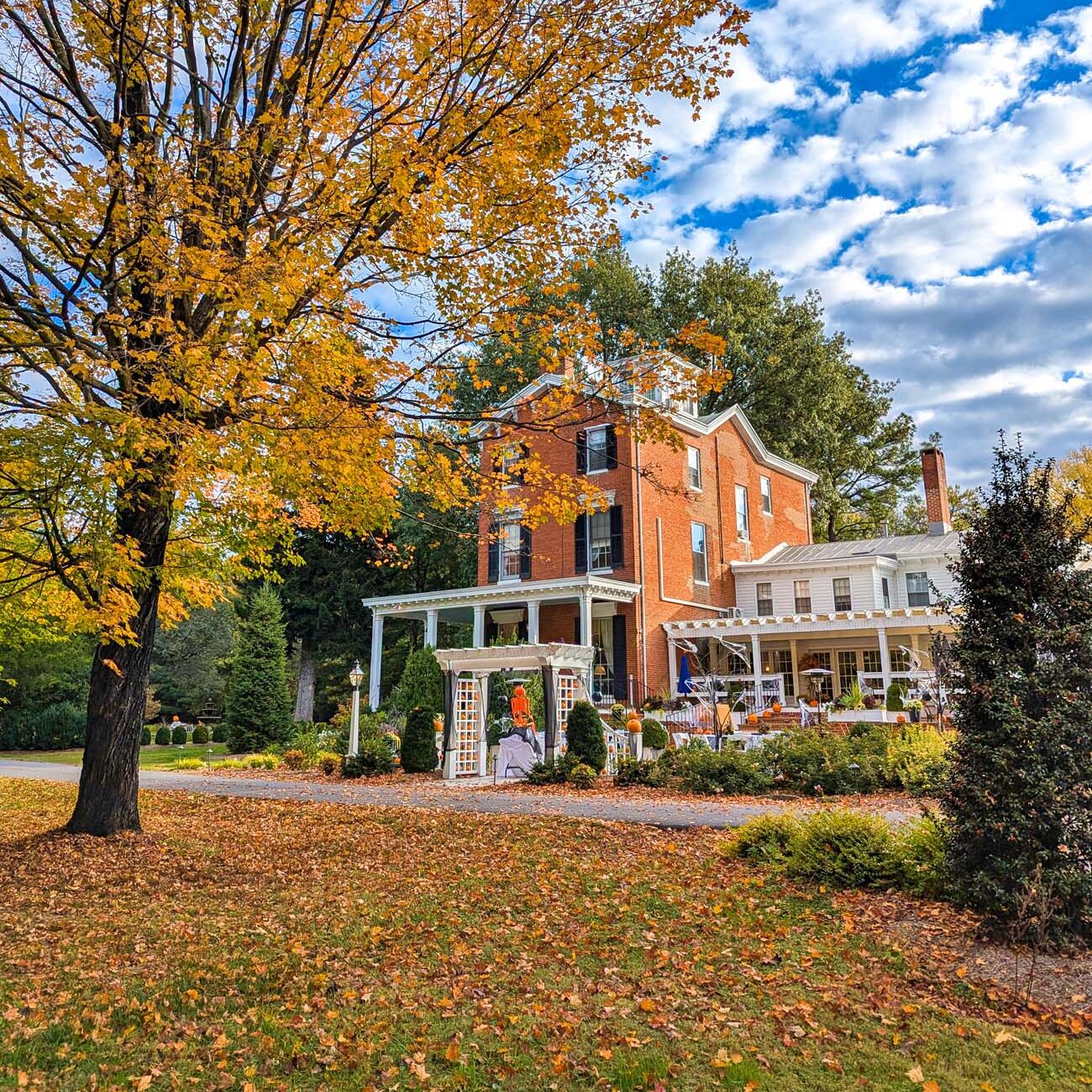 Brampton 1860's three-story red brick Georgian manor house with white columned porch surrounded by vibrant orange and yellow fall foliage under blue sky with puffy clouds