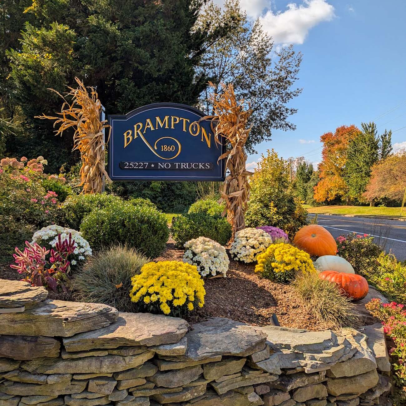 Brampton 1860 blue and gold entrance sign decorated with dried corn stalks, surrounded by chrysanthemums, pumpkins, and ornamental grasses on stone wall