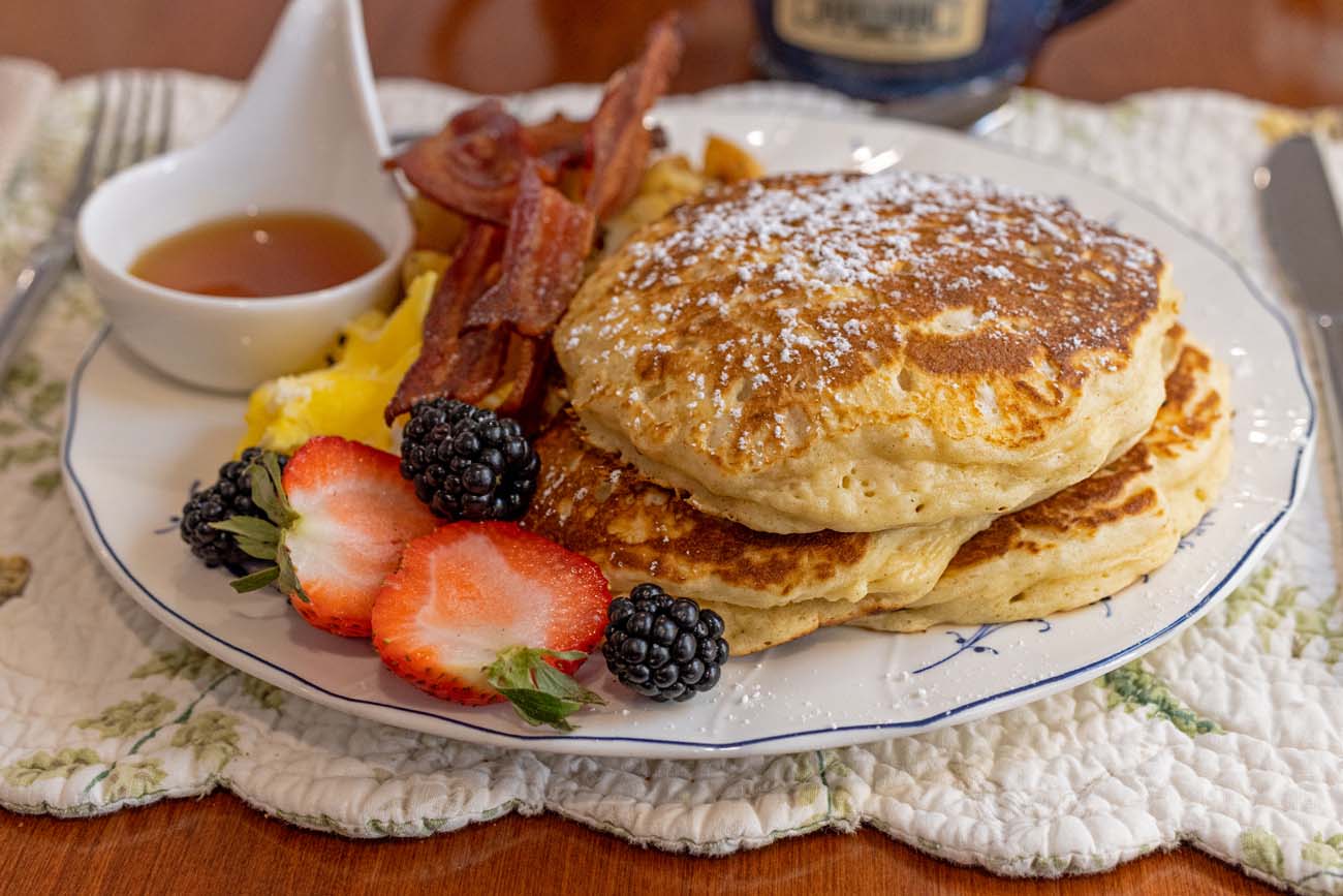 Stack of fluffy pancakes with maple syrup, crispy bacon, scrambled eggs, fresh berries including strawberries and blackberries on white plate with decorative quilt placemat