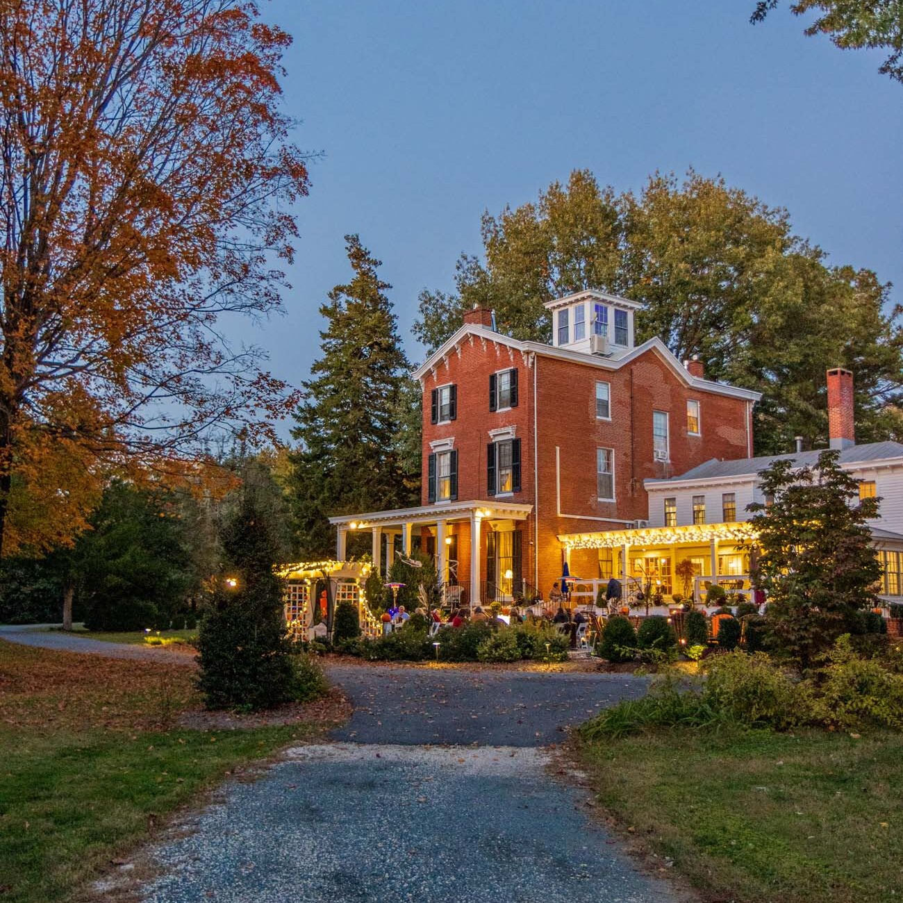 Outdoor evening dining event with guests seated at round tables under string lights, gravel pathway leading to illuminated brick manor house, fall trees surrounding patio area
