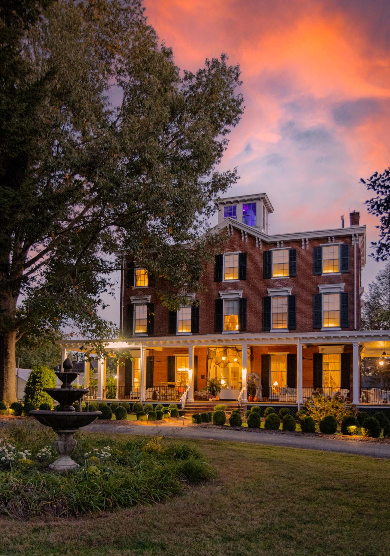 Brampton 1860 manor house at sunset with dramatic pink and orange sky, fountain in foreground, manicured lawn and historic architecture highlighted by warm evening light
