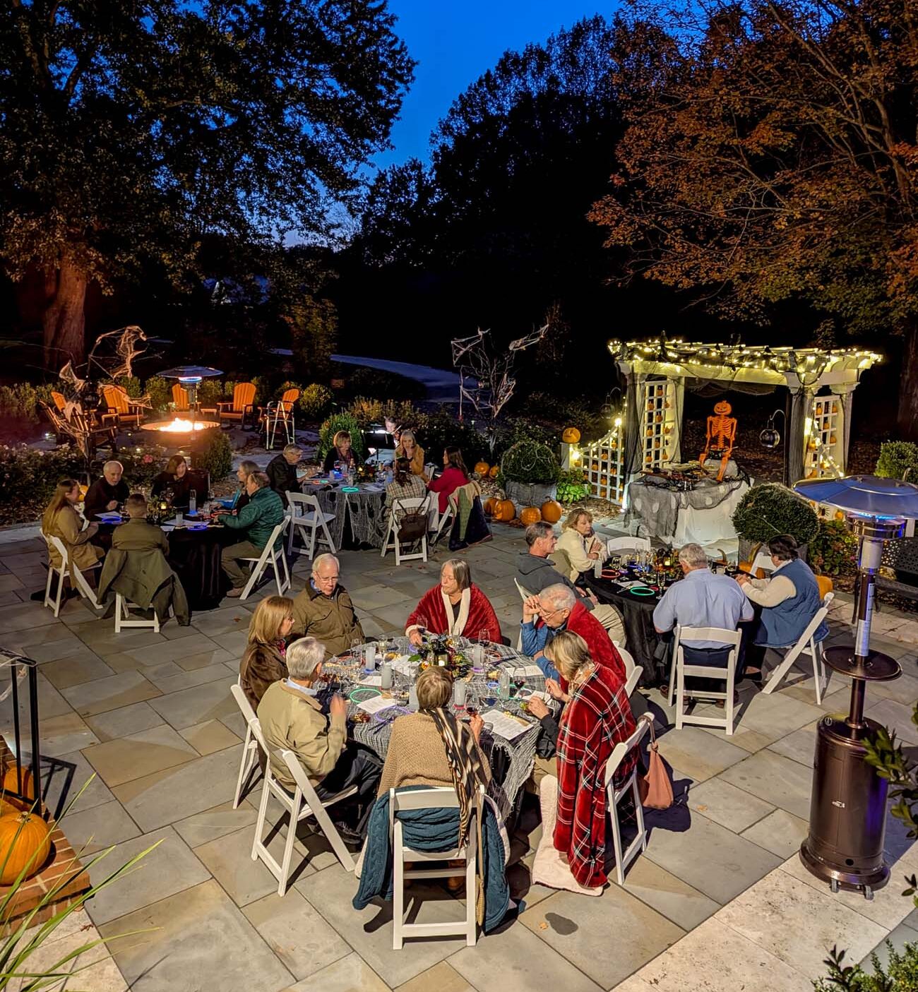 Wide view of evening outdoor dining event with multiple round tables of guests, fire pit area visible, pergola with lights, skeleton decorations at serving station