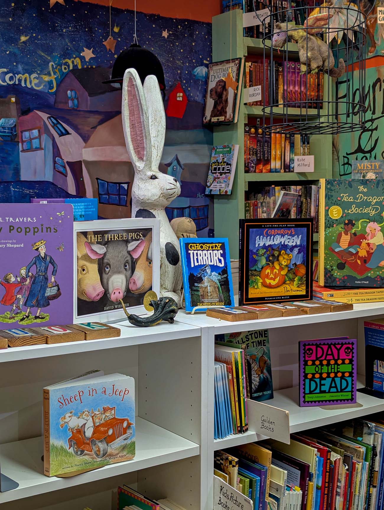 Interior of The Bookplate independent bookstore in Chestertown Maryland showing white bookshelves filled with children's books including The Three Pigs, Ghostly Terrors, and Halloween books, with a large white decorative rabbit statue, whimsical wall mural with stars and houses in background