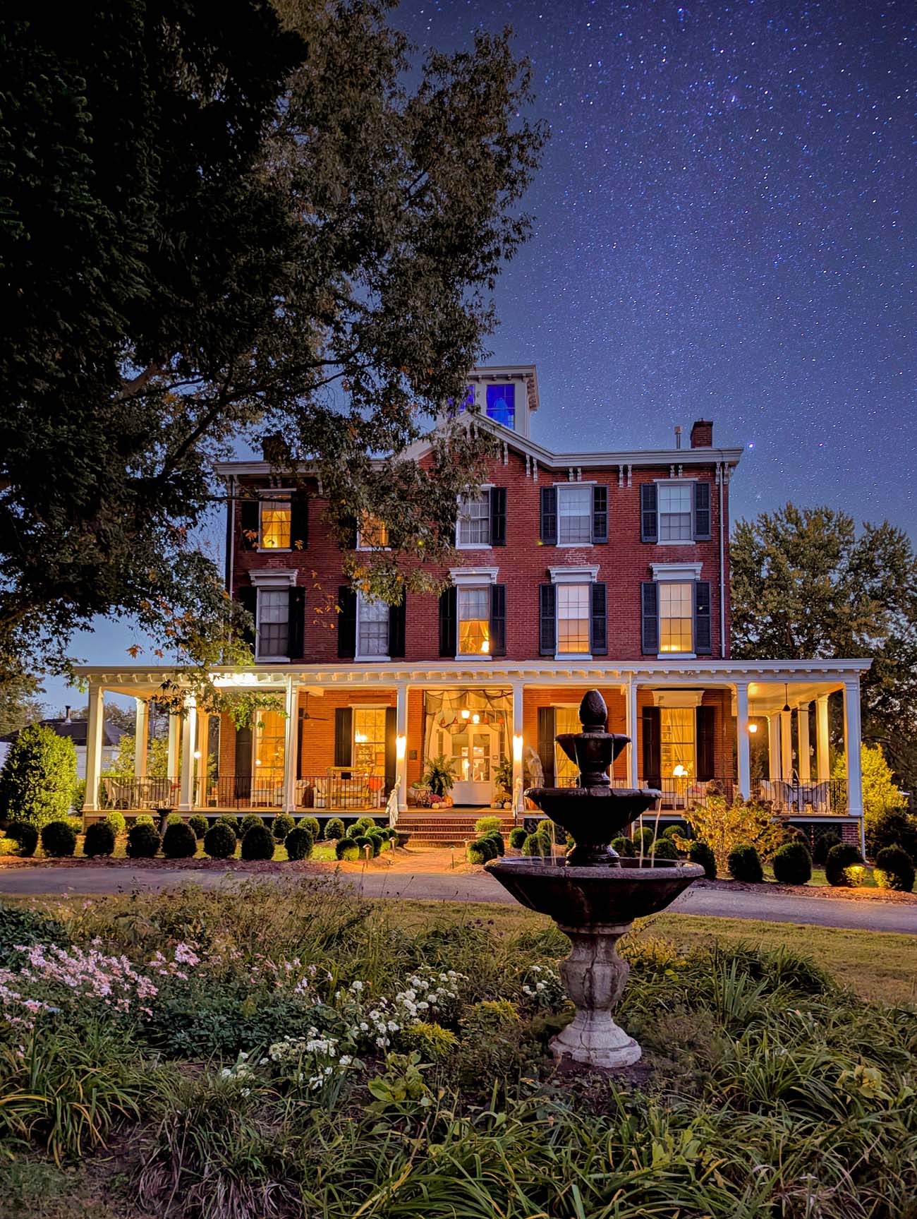 Brampton 1860 historic brick inn at dusk with starry night sky, showing three-story red brick building with white wraparound porch, glowing windows, decorative fountain in manicured garden with colorful flowers, trees on either side