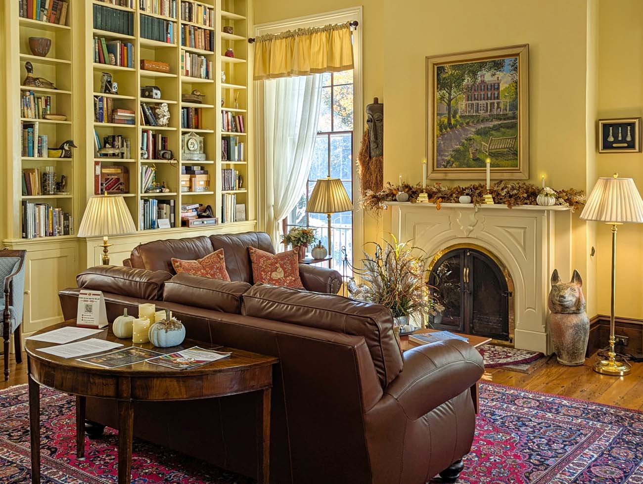 Cozy library room at Brampton 1860 with floor-to-ceiling white built-in bookshelves, brown leather sofa with rust-colored pillows, arched fireplace with white mantel decorated with fall pumpkins and dried flowers, oriental rug, table lamps, and large window with yellow curtains