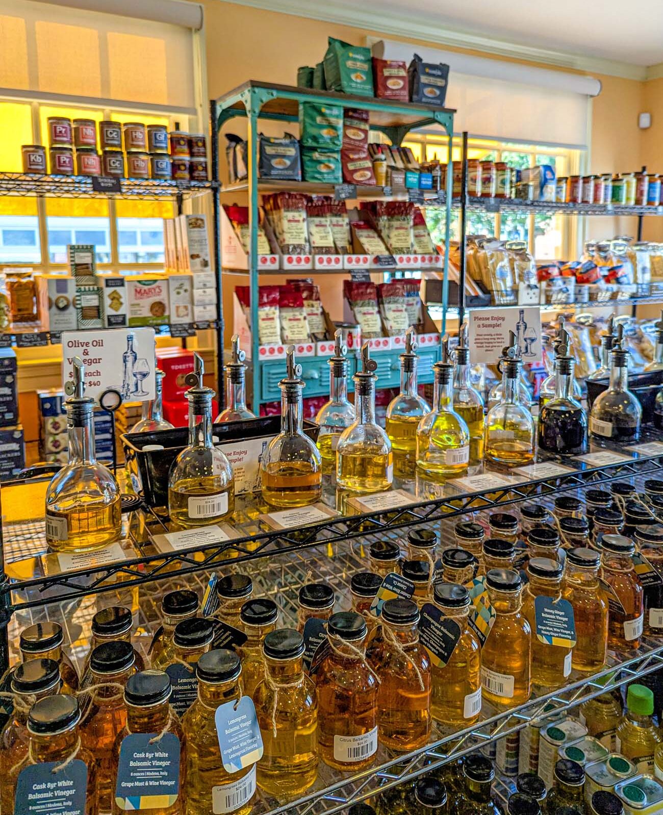 Interior of Chester River Wine & Cheese Co showing wire shelving filled with gourmet products including colorful pasta packages and specialty items on a turquoise shelf unit. In the foreground, a black wire rack displays multiple glass bottles of infused olive oils with pour spouts for sampling, alongside rows of small jars containing flavored honey in golden and amber hues. Additional shelves in the background hold various gourmet food products. Warm yellow walls and natural light from windows create an inviting atmosphere