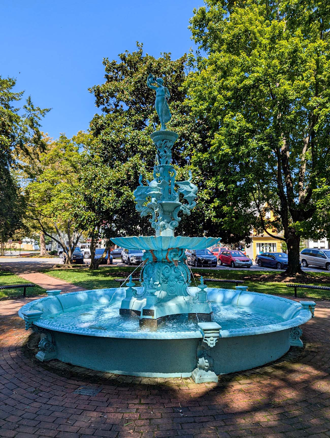 Ornate Victorian-era fountain painted in turquoise blue located in a town square in Chestertown, Maryland. The multi-tiered cast iron fountain features elaborate scrollwork, decorative flourishes, and a female figure statue at the top. Water flows from multiple levels into a circular basin below. The fountain sits on brick pavement surrounded by a green lawn, with mature trees providing dappled shade. Historic buildings and parked cars are visible in the background under a clear blue sky.
