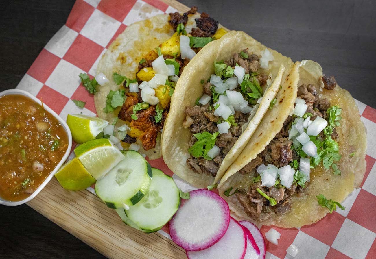 Three authentic Mexican street tacos on warm corn tortillas with seasoned meat, diced onions and fresh cilantro on red and white checkered paper, served with cucumber slices, radish, lime wedges and small bowl of salsa verde on wooden board