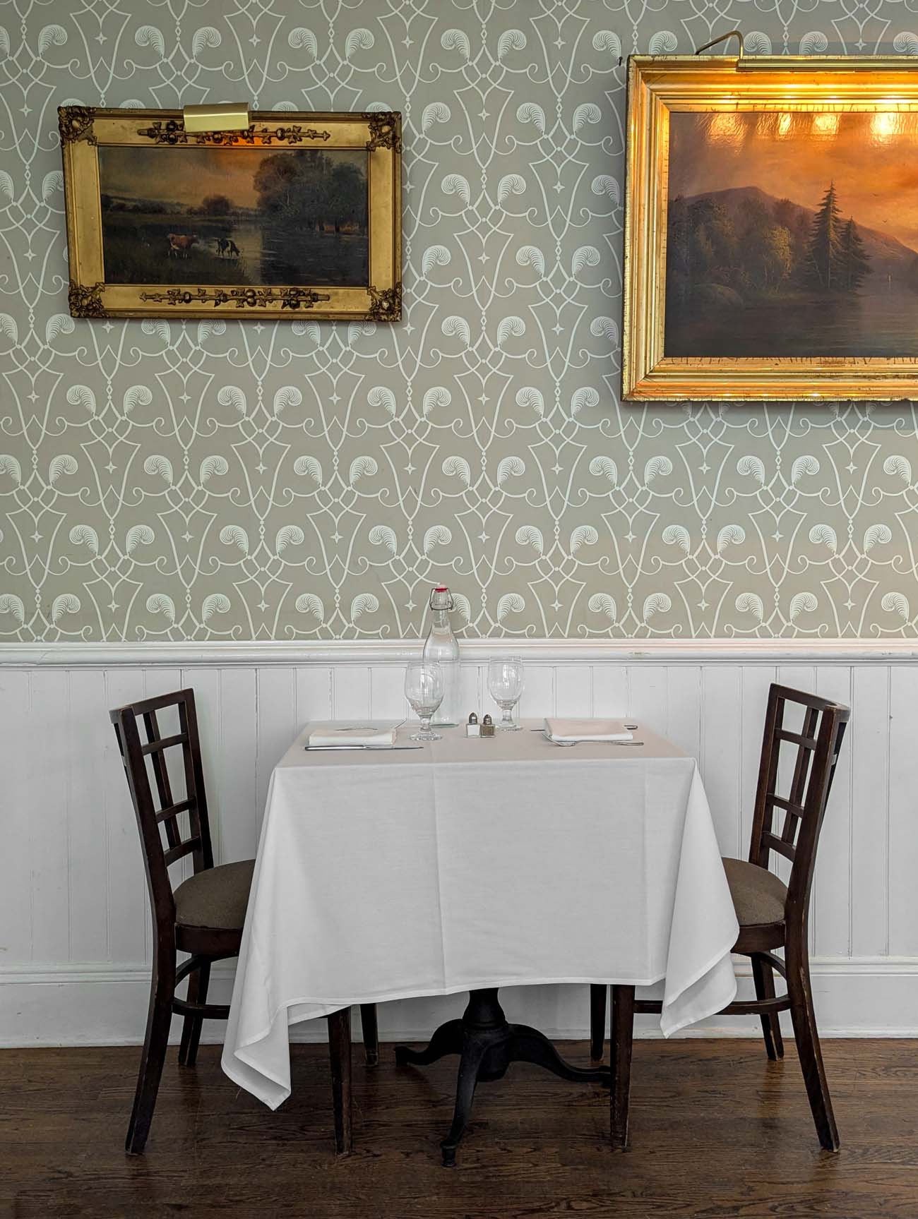 Elegant intimate table for two in historic Delmonico Room restaurant with white tablecloth, vintage pastoral paintings on sage green patterned wallpaper and white wainscoting at Michelin Key Hotel Fauchere Milford Pennsylvania