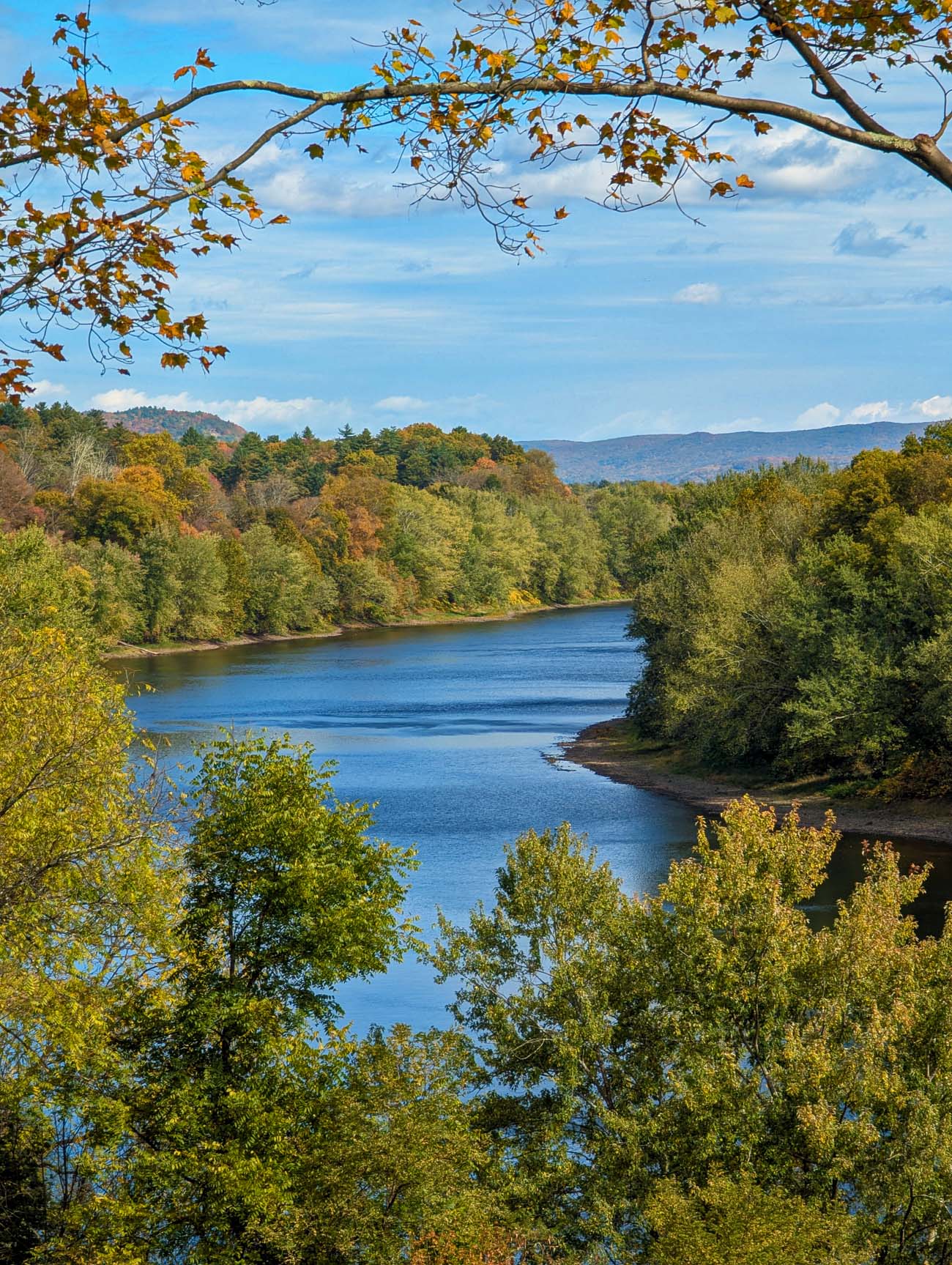 Peaceful Delaware River shoreline at Milford Beach Pennsylvania in autumn showing rocky pebble beach, calm blue water, green steel pedestrian truss bridge, forested hillsides with orange and yellow fall foliage, clear blue sky, northern gateway to 37-mile McDade Recreational Trail in Delaware Water Gap National Recreation Area Pike County
