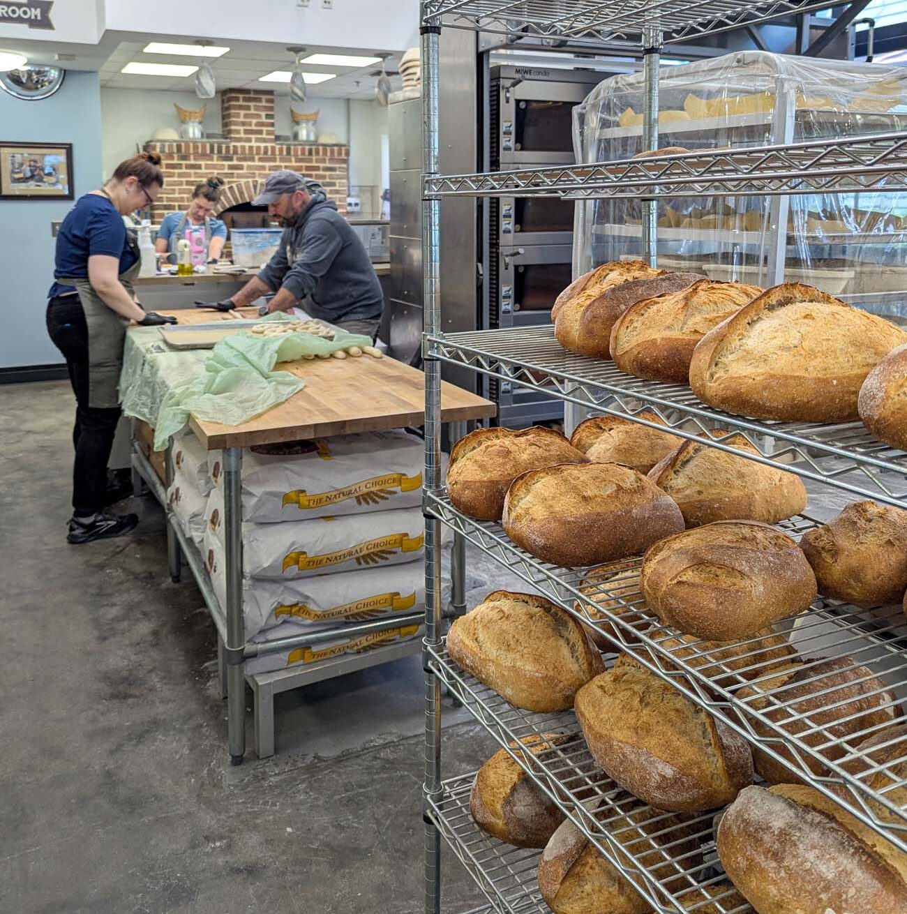 Behind-the-scenes view of Modern Stoneage Kitchen bakery showing professional bakers working at a large wooden table preparing dough. Wire shelving racks in the foreground display dozens of freshly baked artisan bread loaves with golden-brown crusts dusted with flour. A brick oven is visible in the background along with stainless steel commercial baking equipment. The industrial kitchen features polished concrete floors and bright overhead lighting.