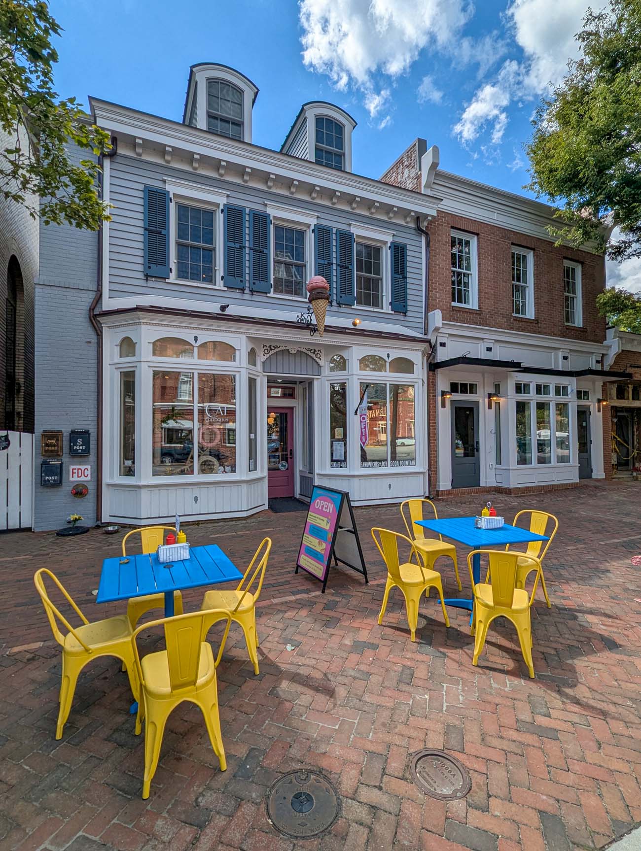 Stam's Luncheonette exterior showing historic white two-story building with black shutters, curved bay windows on first floor, red door entrance, bright yellow metal bistro tables and chairs on brick plaza, ice cream cone sign, blue sky