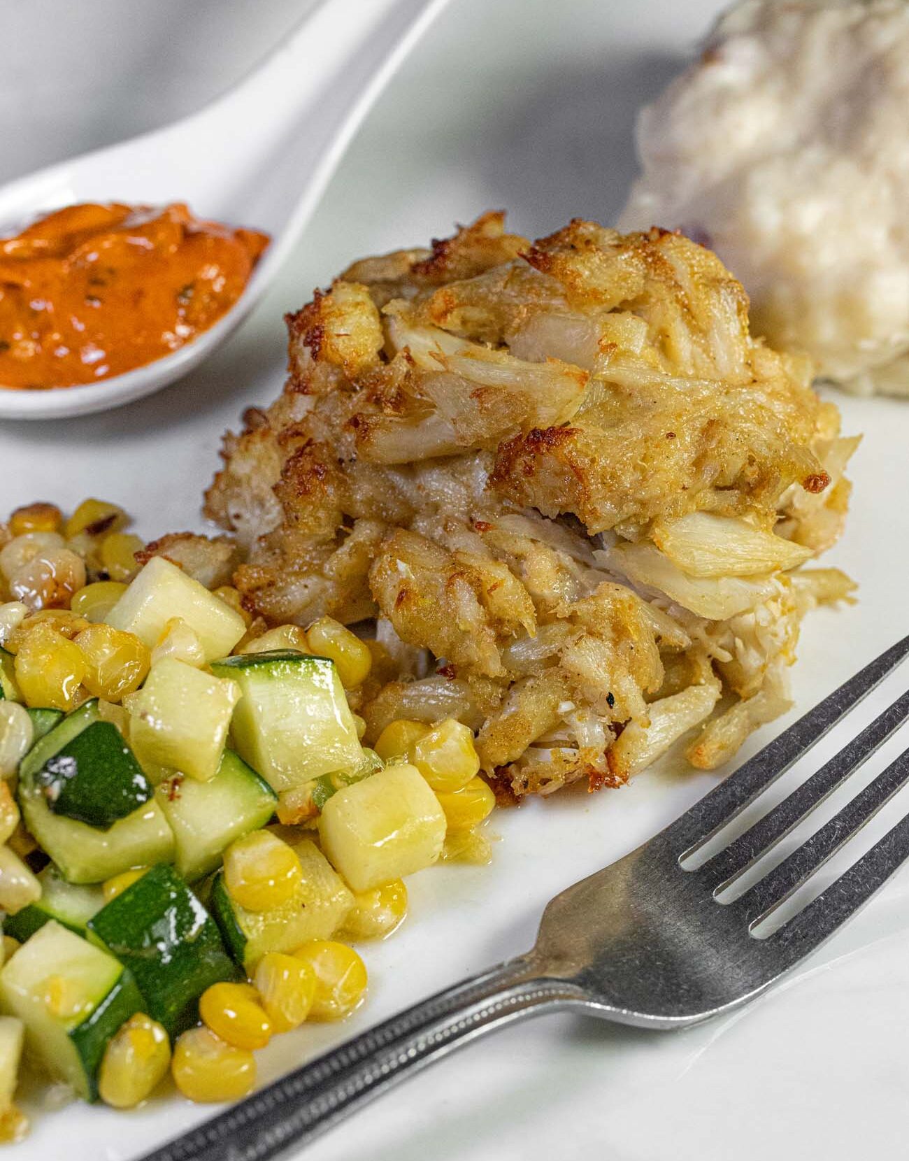 lose-up of a golden-brown jumbo lump crab cake with crispy edges on a white plate, accompanied by a side of sautéed zucchini and corn succotash. A ramekin of orange remoulade sauce and what appears to be coleslaw are visible in the background. A silver fork rests on the right side of the plate. The crab cake shows large chunks of premium crab meat with minimal filler and a perfectly seared crust.