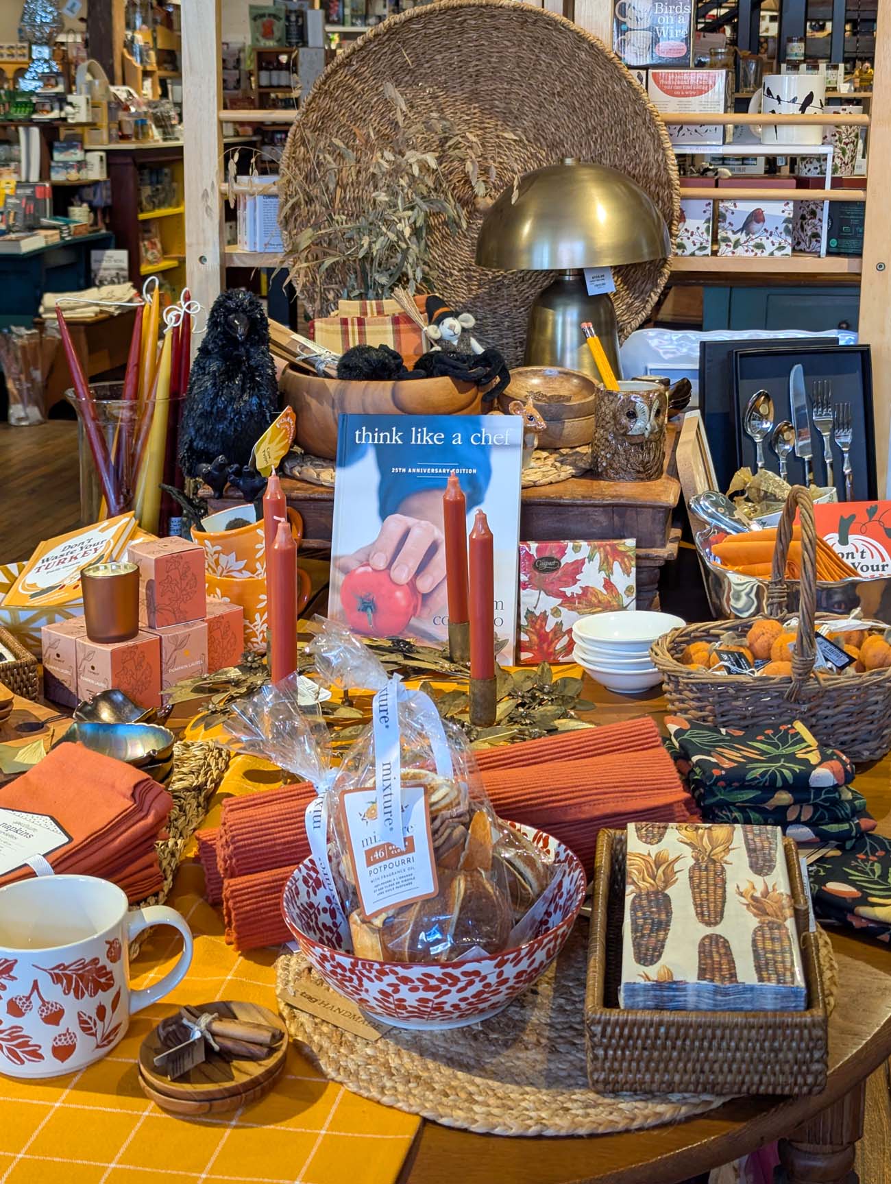 Twigs and Teacups gift shop interior display showing eclectic home goods including woven basket, black poodle figurines in wooden bowl, gold dome lamp, flatware set, think like a chef cookbook, decorative candles, orange placemats, patterned mugs, colorful linens and kitchen accessories arranged on counter with bookshelves visible in background