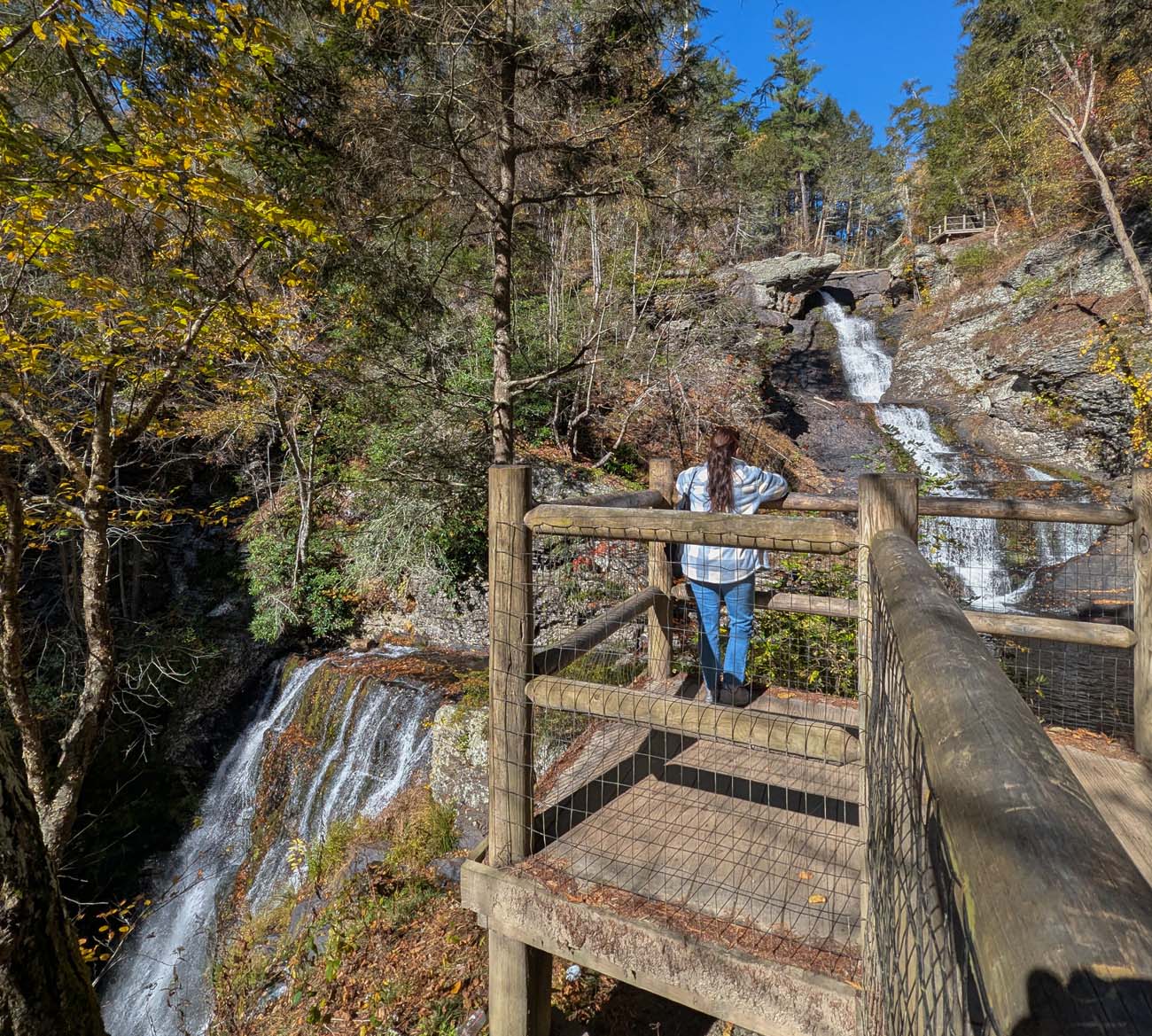 Woman viewing Pennsylvania's tallest waterfall Raymondskill Falls from wooden observation platform showing three tiers of cascading water at 178 feet surrounded by autumn forest foliage in Delaware Water Gap National Recreation Area near Milford PA